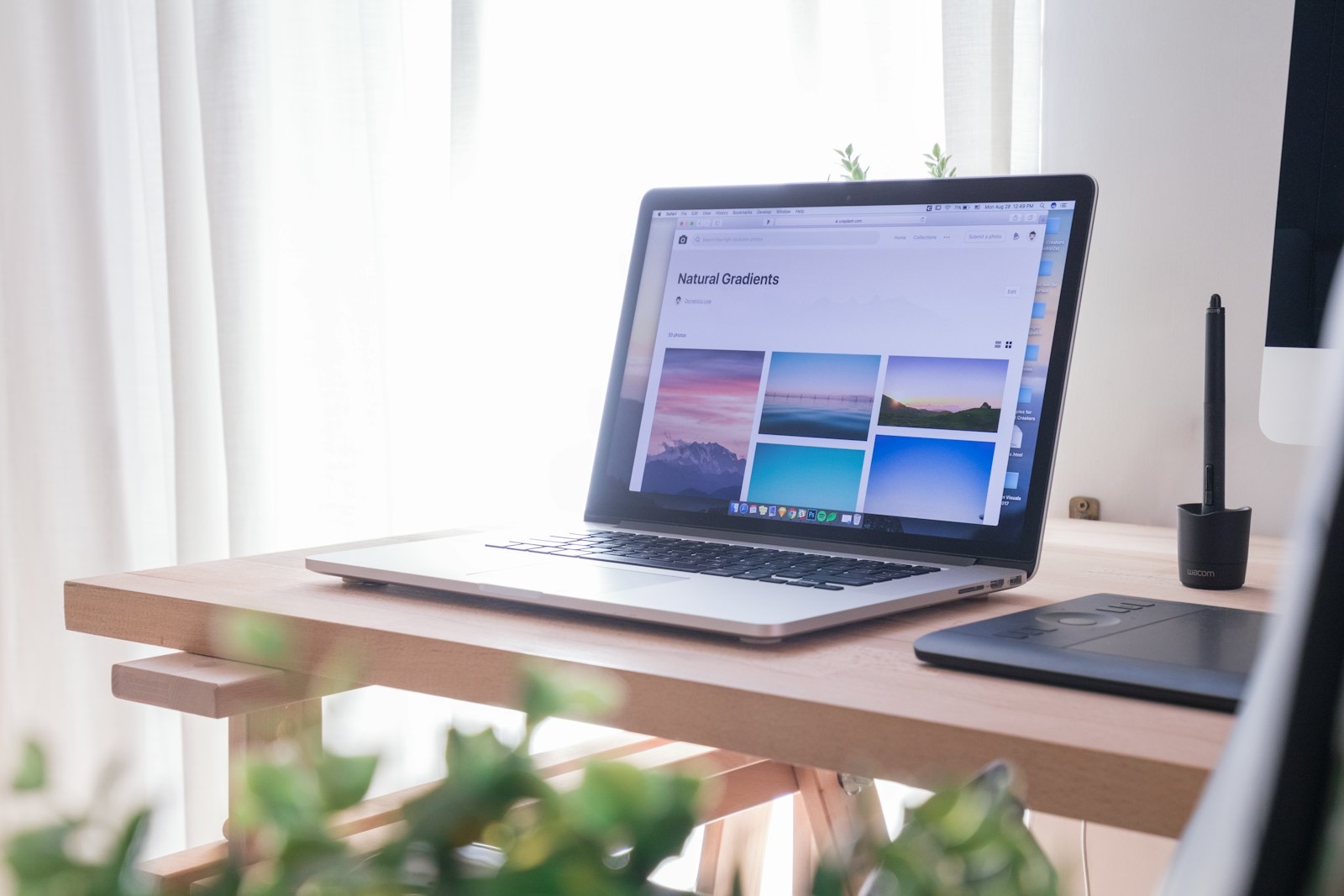 An open laptop on a light wooden desk by a bright window, with a drawing tablet beside it and a plant in the foreground.