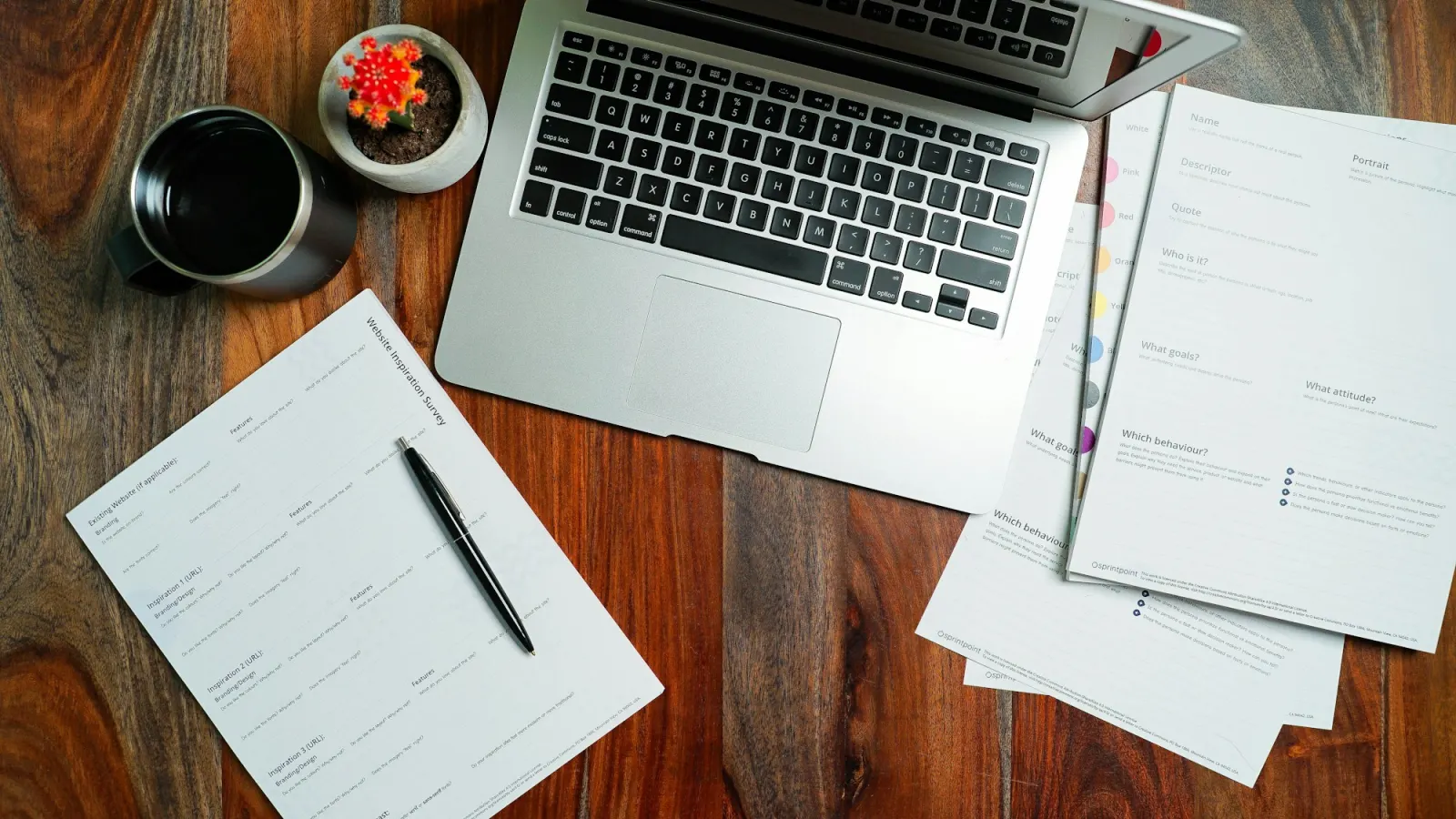 Top-down view of a laptop, a coffee mug and printed website planning worksheets with a pen, on a wooden desk.