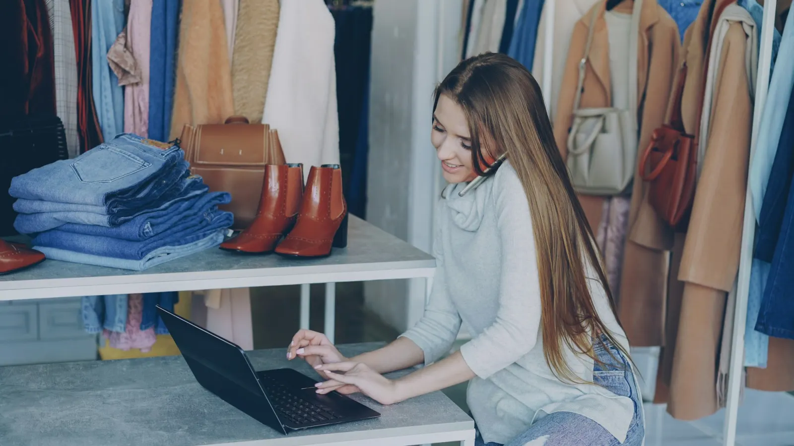 A clothing shop owner on the phone while typing on a laptop at her shop counter, with jeans, boots and coats on the rails behind her.