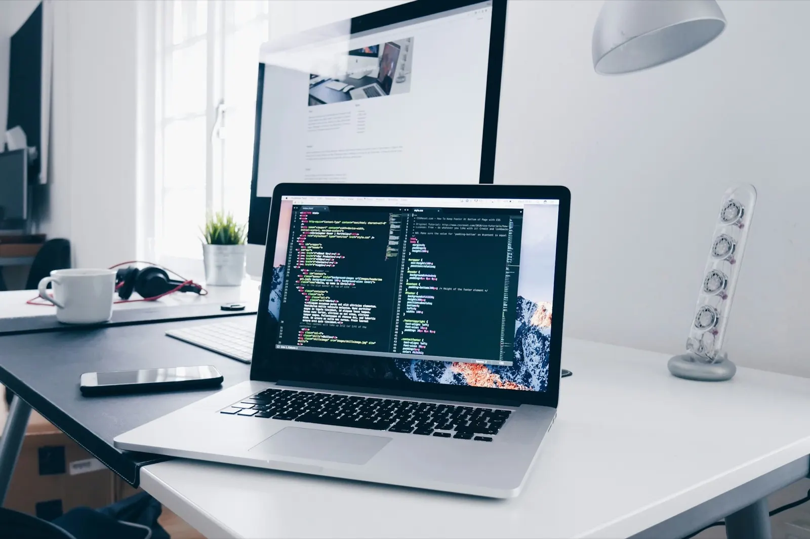 A laptop showing HTML and CSS code on a white desk, with a large monitor, keyboard, phone and coffee mug behind it.
